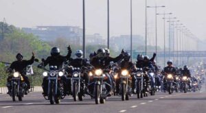 Image of a motorcycle rider and pillion passenger not wearing helmets on a busy road in India. The rider is holding a helmet in hand, highlighting the common practice of not using helmets for safety but for avoiding fines. In the background, other vehicles can be seen on the road, showcasing typical Indian traffic conditions.